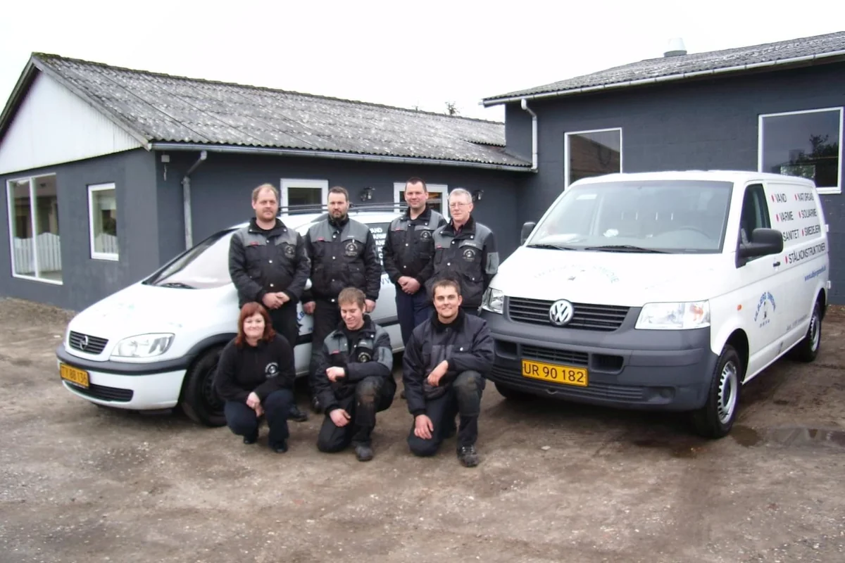 Medarbejdere fra Ulbjerg VVS foran virksomhedens bygning sammen med to servicebiler.